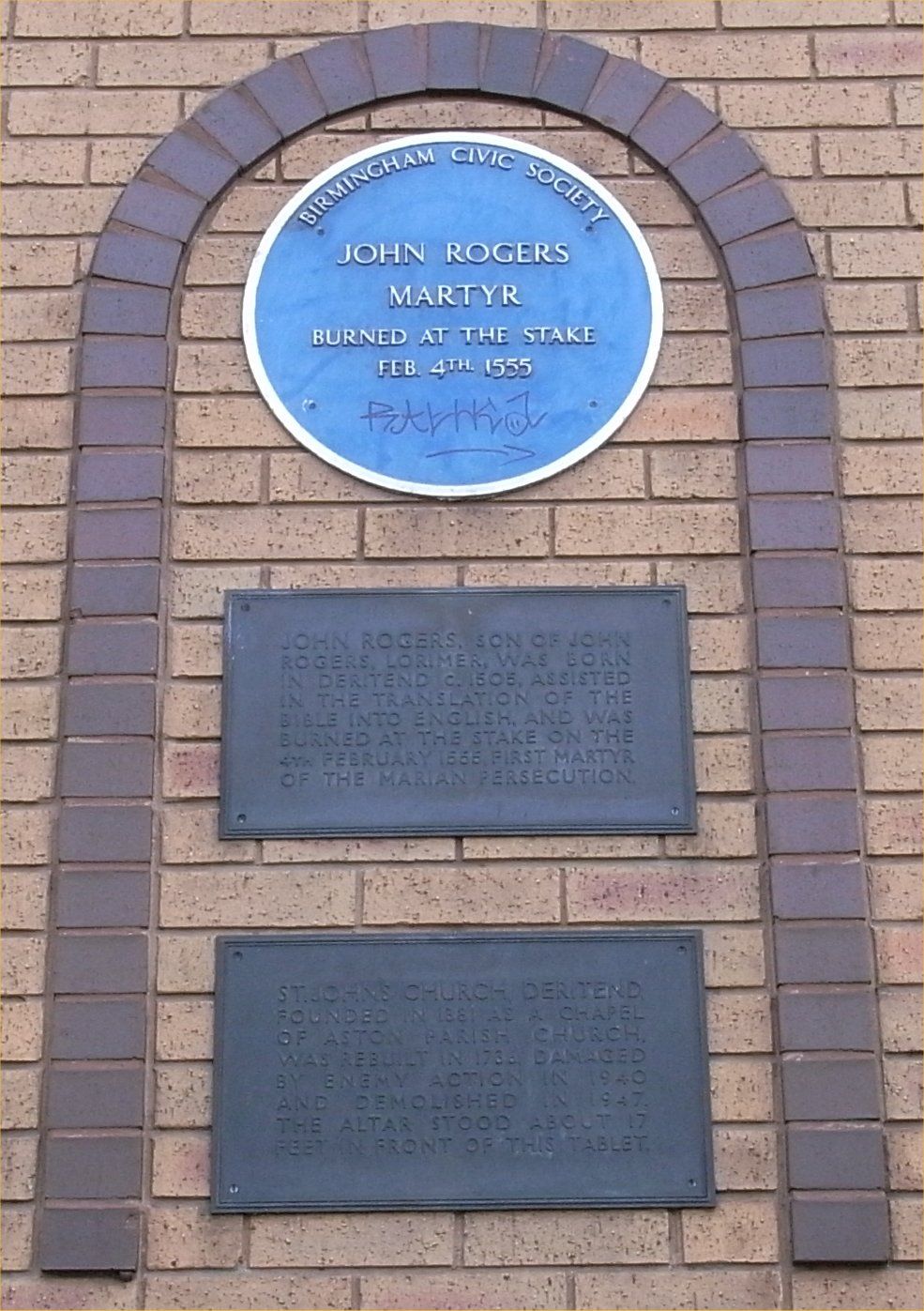 A blue plaque to John Rogers above two bronze tablets set in a brick arch.