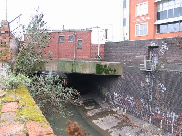 A river in a brick channel running into a culvert under buildings.