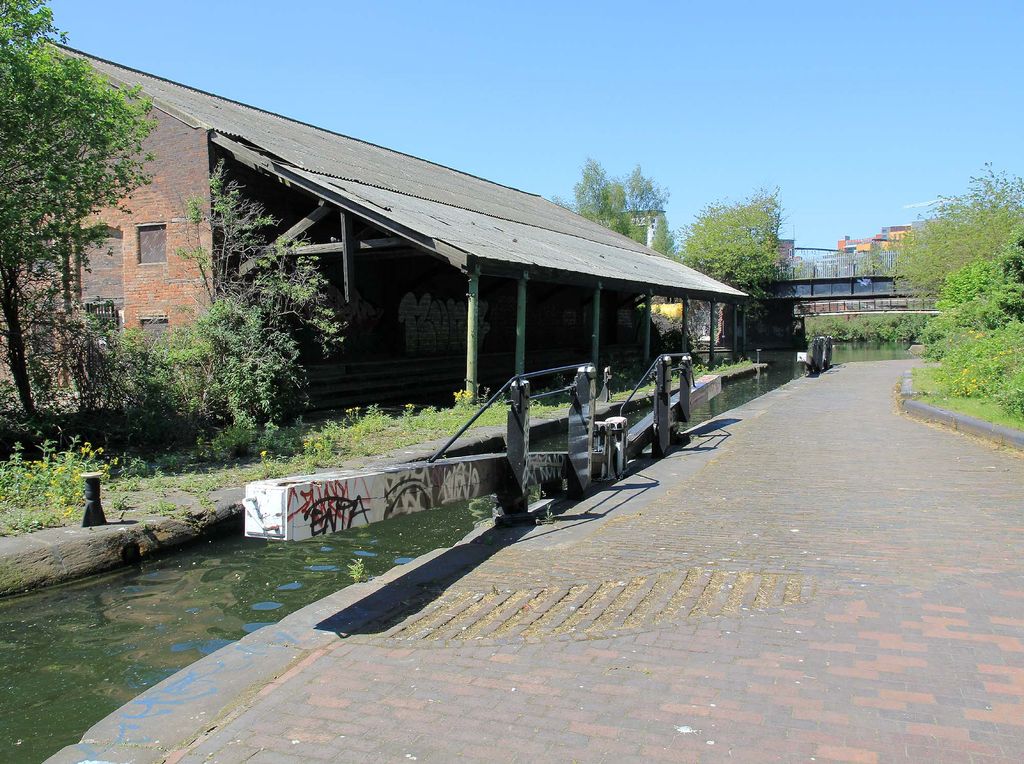 A canal stop lock with black-and-white balance beams beside a long brick warehouse whose roof overhangs the water on iron columns.