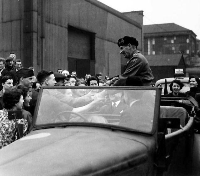 General Montgomery in a beret stands in an open army car as a crowd of factory workers, many of them women, reach in to shake his hand beside works buildings.