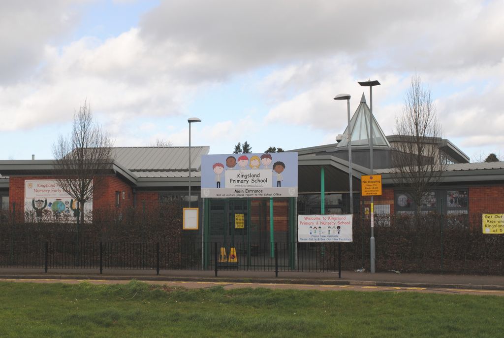 Single-storey brick school buildings with a modern glazed pyramid roof, railings and a school sign, grass in front