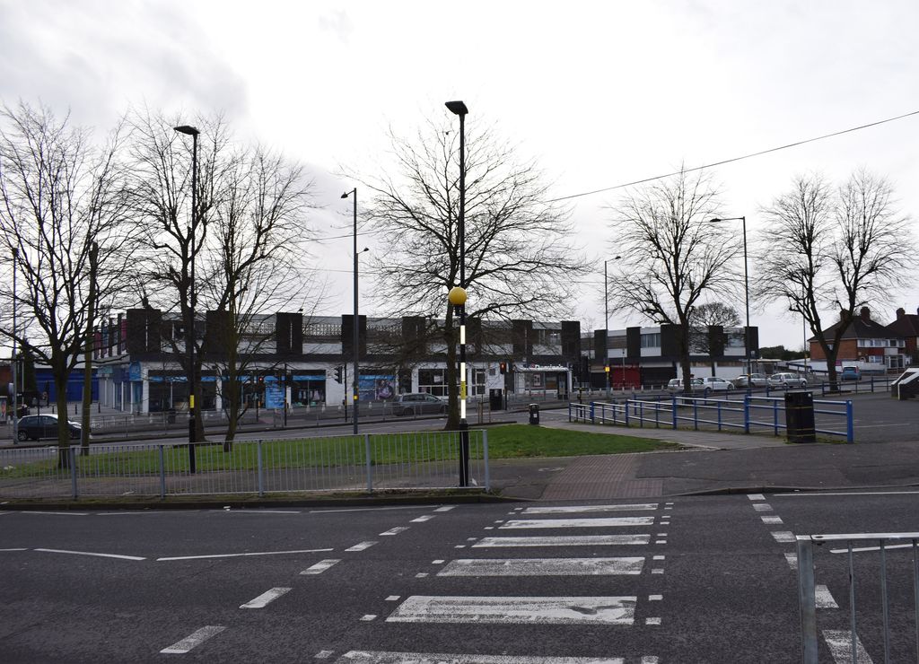 Row of flat-roofed 1960s precinct shops behind bare trees across the Circle from Kettlehouse Road, with a pedestrian crossing in front