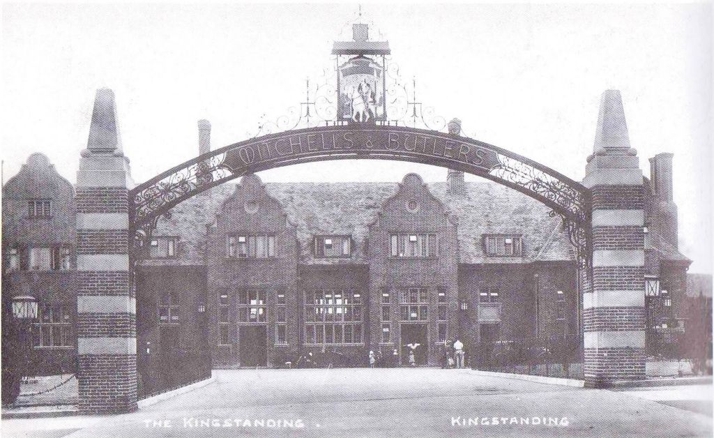 The Kingstanding pub behind a tall iron arch lettered Mitchells & Butlers, with a hanging sign of a rider on a white horse, striped brick piers and Dutch gables
