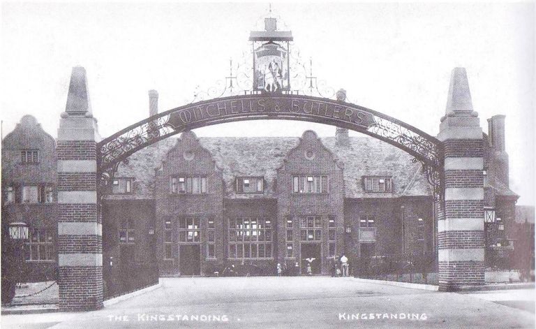 The Kingstanding pub behind a tall iron arch lettered Mitchells & Butlers, with a hanging sign of a rider on a white horse, striped brick piers and Dutch gables