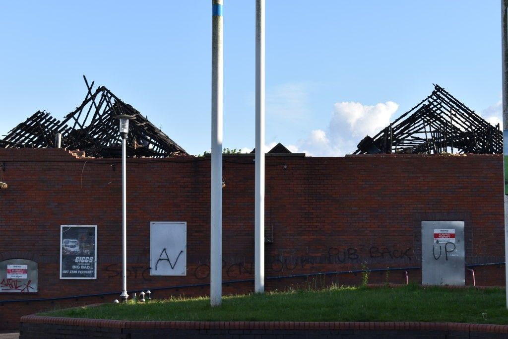 Burnt-out roof timbers above the brick walls of the former Kingstanding pub, with flagpoles and graffiti in the foreground, May 2019