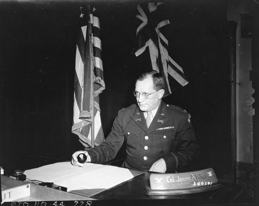 US Army colonel in uniform and glasses seated at a desk with a nameplate reading Col. James A. Kilian, the Stars and Stripes and Union Jack behind him