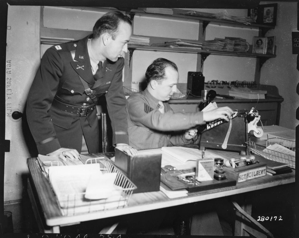 Two American soldiers in an office: a captain leaning over a desk while a staff sergeant works an adding machine, a Sgt Gilbert nameplate on the desk