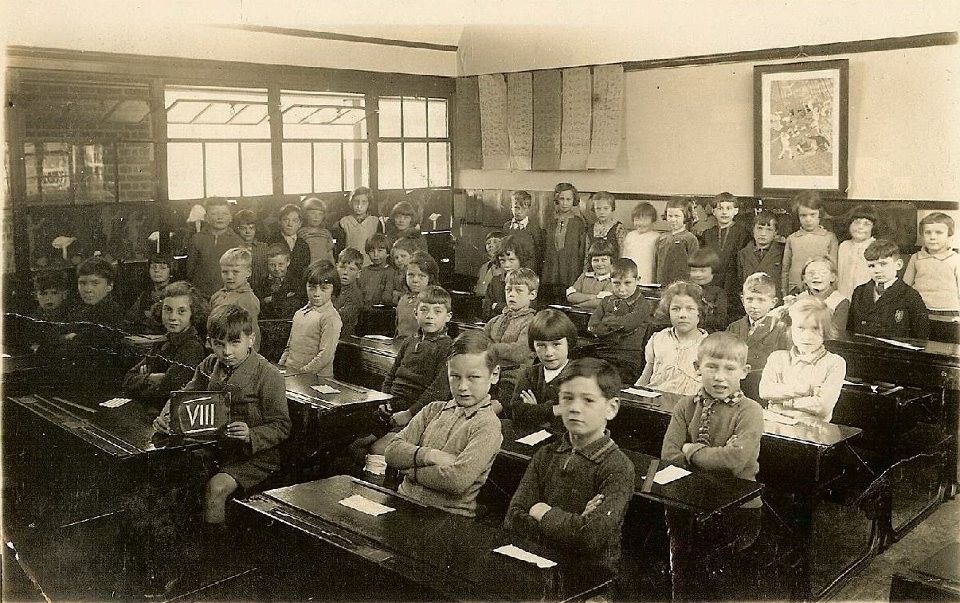 Black-and-white class photograph of about forty children sitting at wooden double desks in a classroom, a boy in front holding a slate marked VIII