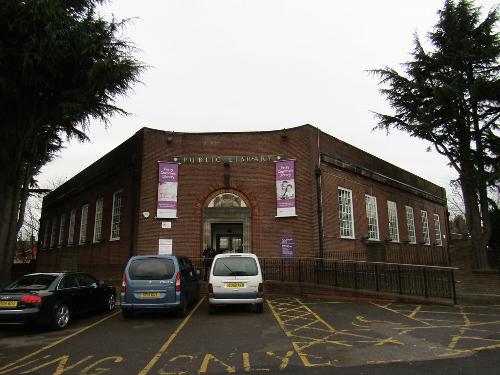 Single-storey brick library with a curved corner, 'PUBLIC LIBRARY' lettering, an arched stone entrance and purple banners, cars parked in front
