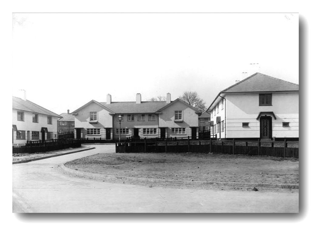White-rendered estate houses around a curving road and a large grass island, with a gas lamp and wooden fences