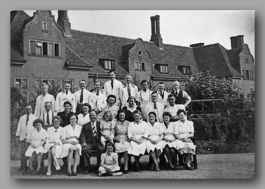 A group of about thirty men, women and children, many in white coats and aprons, posed on a lawn in front of a building with Dutch gables