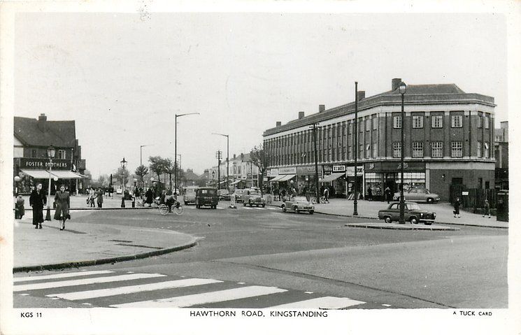 Black-and-white postcard of Hawthorn Road, Kingstanding, around 1950: shoppers and cyclists by a large shopping block, parked cars, and Foster Brothers on the corner